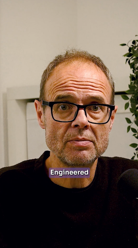 A close-up portrait of a man with glasses and a short beard, wearing a black shirt, with a purple label reading 'Engineered' over the chest. A plant is visible on the right side of the background.